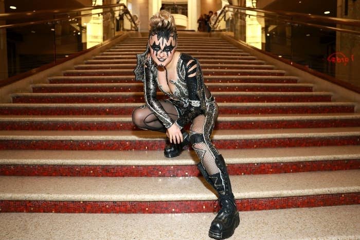 JoJo in a detailed Kiss-inspired outfit crouching on a staircase. The costume has metallic elements, fishnet stockings, and dark platform boots