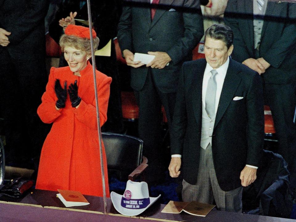 Nancy in a bright red coat and matching hat and black gloves clapping next to her husband.