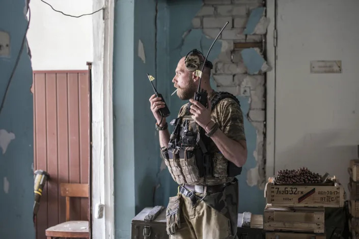 A Ukrainian soldier with a cigarette in his mouth, looks out of a decrepit building, holding a radio in each hand. 