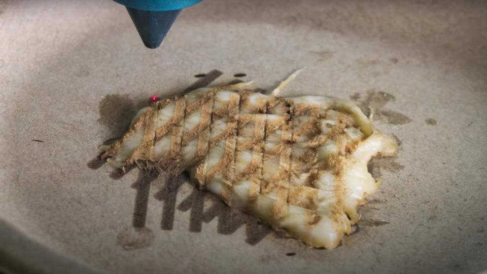 A laser cooking up a piece of flat, 3D-printed chicken on top of a beige surface. 