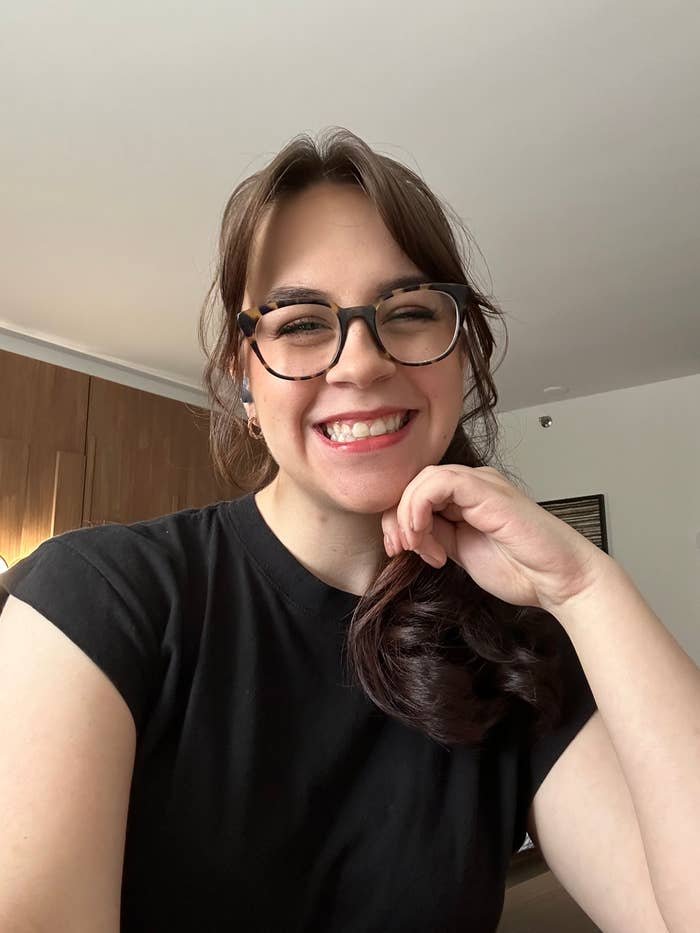 A woman with glasses and a black shirt smiles warmly, resting her chin on her hand. She appears in an indoor setting with wooden panels on the wall