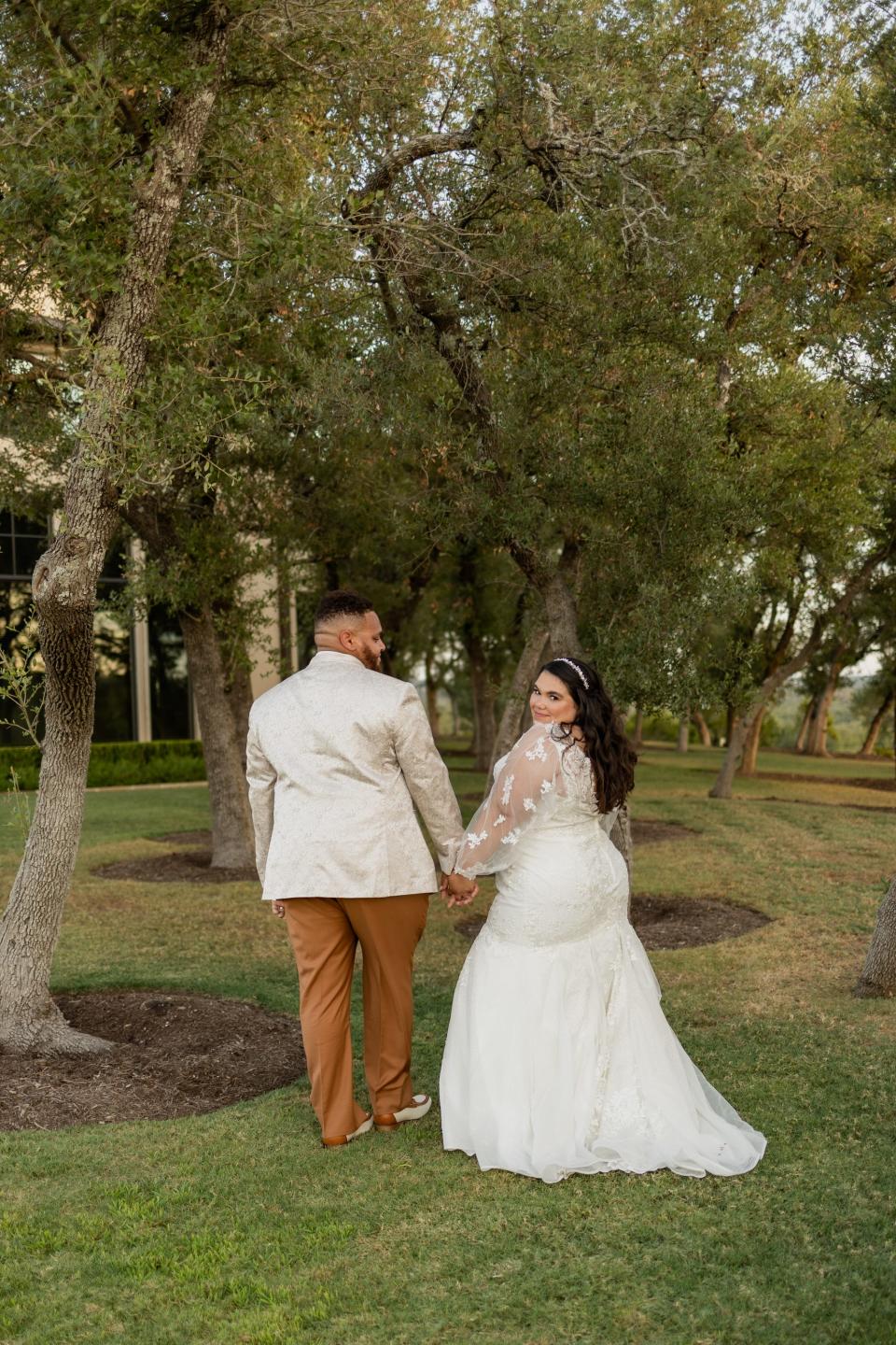 A bride looks over her shoulder as she hold hands with her groom in a field.