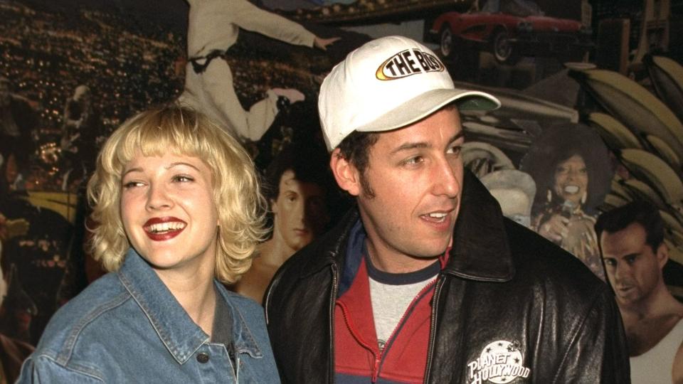 drew barrymore in a denim jacket stands next to adam sandler with her hand on his chest, she smiles and looks left of the camera, sandler looks right of the camera, he wears a black leather jacket with a planet hollywood logo a white baseball cap and a red and blue zipup jacket