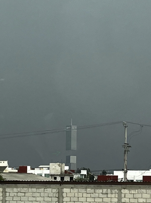 A tall, narrow structure with the word "OMA" at the top is seen from a distance amidst a gray sky and urban foreground