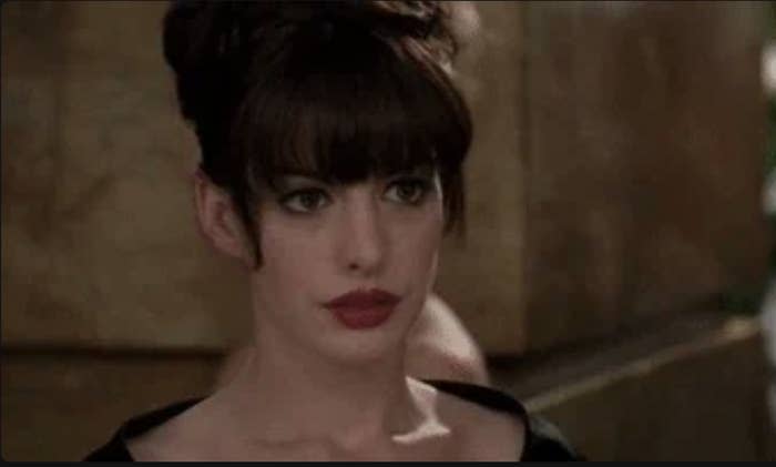 Anne Hathaway with an updo hairstyle and bangs, wearing elegant makeup and a black outfit, looks contemplative