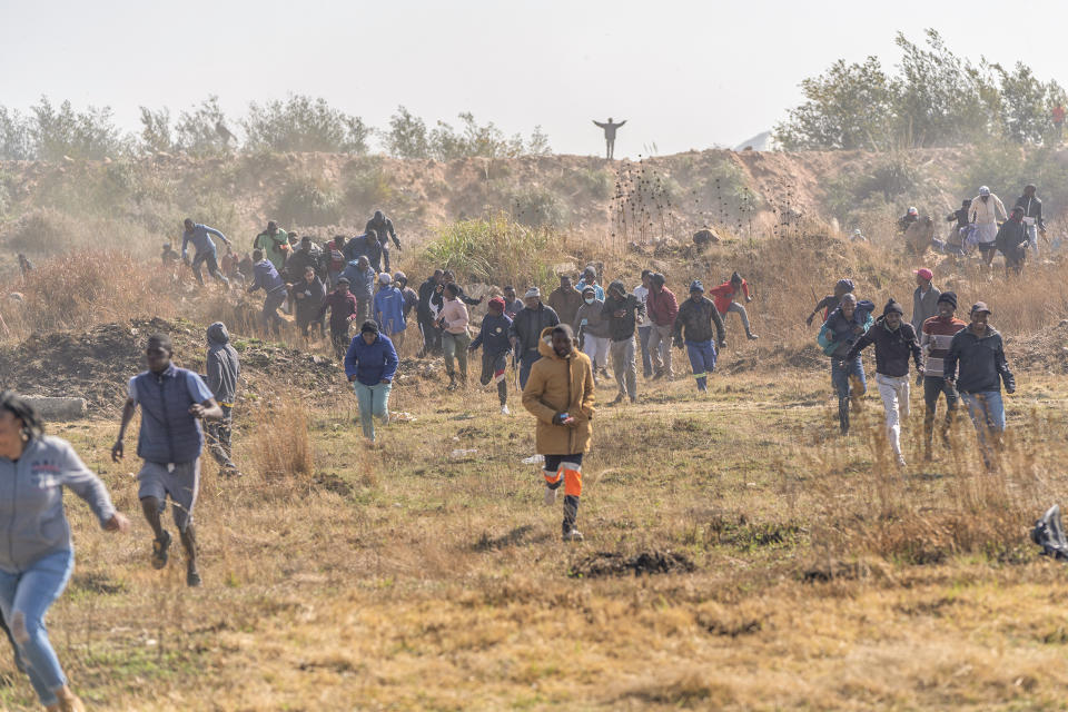 People run from tear gas and stunt grenades launched from a police helicopter in the South African city of Krugersdorp Thursday Aug. 4, 2022. Community members in the assaulted suspected illegal miners and set fire to their camps on Thursday in an outpouring of anger following the alleged gang rapes of eight women by miners last week. Residents of Krugersdorp's Kagiso township also barricaded roads with rocks and burning tires during a planned protest.(AP Photo/ Shiraaz Mohamed)