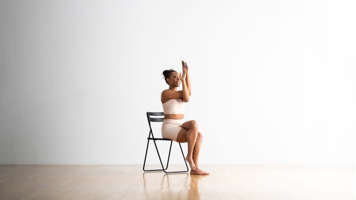 Woman practices Garudasana, Eagle Pose,on a chair. She wears white yoga shorts and top and sits against a white background