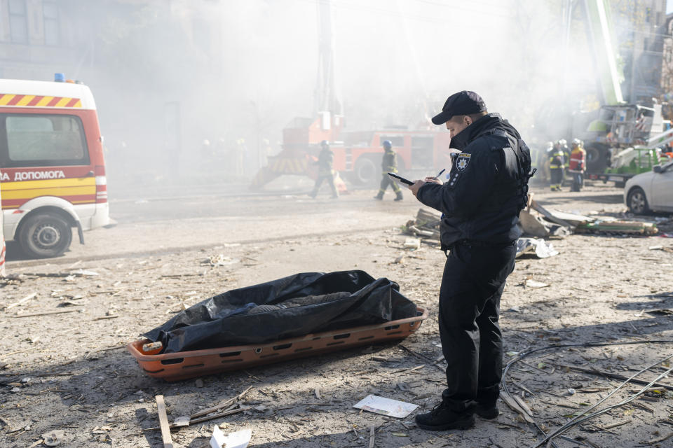 A police officer writes on a pad, looking at a body bag placed in a long container on a debris-strewn street, with an emergency vehicle in the background.