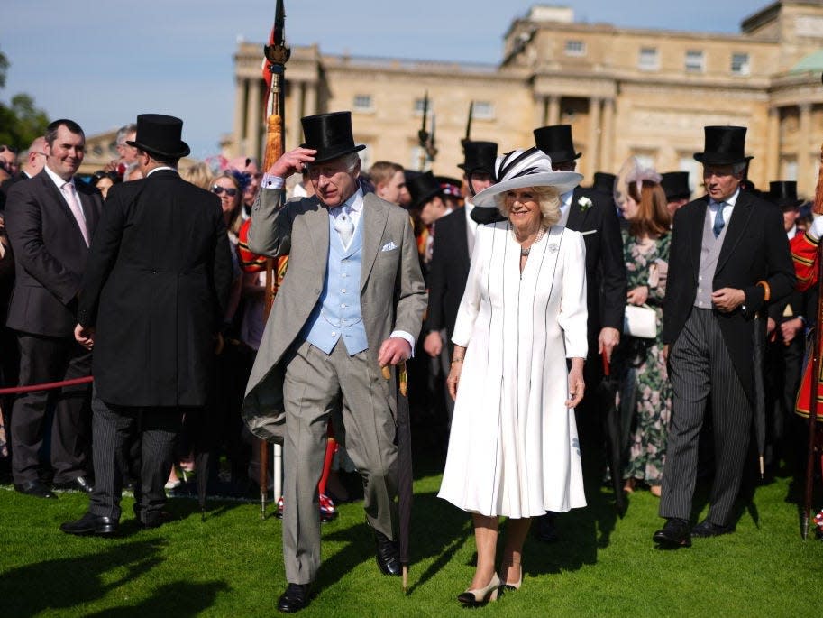 King Charles III wearing a three-piece suit and a top hat and Queen Camilla wearing a white dress and white fascinator in the gardens of Buckingham Palace.