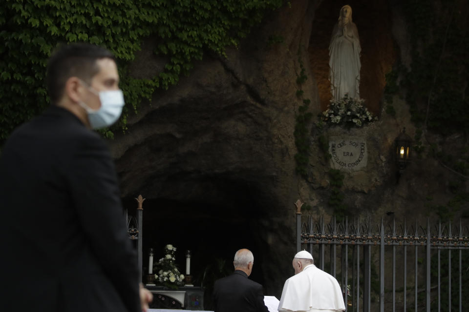 A priest wears face mask to prevent the spread of COVID-19, as Pope Francis prays during a rosary in Vatican gardens Saturday, May 30, 2020. In background is a reconstruction of the Lourdes' grotto in southern France, where the Virgin Mary is alleged to have appeared eighteen times to Bernadette Soubirous in 1858. Pope Francis is reciting a special prayer for the end of the coronavirus pandemic surrounded by a representative sampling of people on the front lines in his biggest post-lockdown gathering to date. (AP Photo/Alessandra Tarantino, pool)