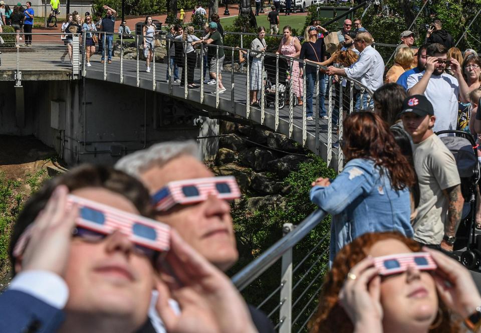 People fill the Falls Park bridge near Barnes Elliott, left, State Rep. Jason Elliott, and Tabitha Bone of Greenville, taking a break from work to view the solar eclipse at the peak time in Greenville, S.C. Monday, April 8, 2024.