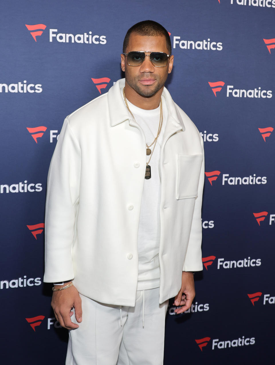 Russell Wilson at a Fanatics event, wearing a white jacket and pants, with sunglasses, and layered gold necklaces