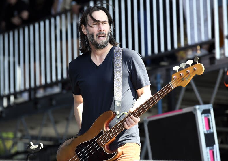 Keanu Reeves, hair long and unshaven, holds bass guitar while wearing a black t-shirt, performing on stage with Dogstar.