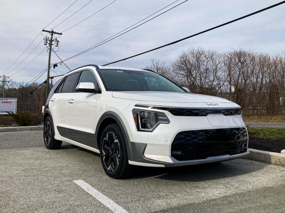 A white Kia Niro EV electric SUV is seen in a parking lot against an overcast sky, with a Tesla charging station in the foreground.