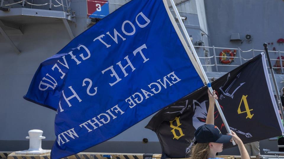 Family of Sailors aboard the Navy destroyer Bulkeley wave the ship’s battle flag as it prepared to depart Naval Station Norfolk, Virginia, and head to its new home port in Rota, Spain, in August 2022. (Mass Communication Specialist 1st Class Theodore Green/Navy)