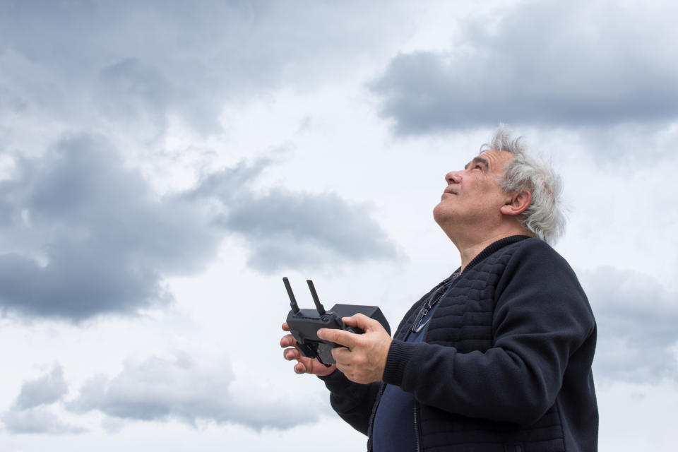 An older man with gray hair is looking at the sky while holding a drone controller. Cloudy sky background