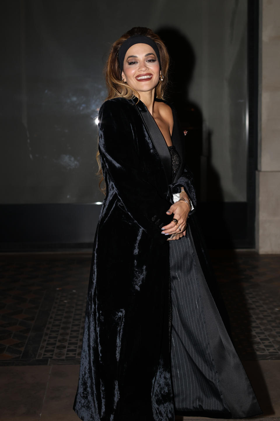 Rita Ora smiles in Paris while wearing a black coat, black dress, black pumps, and thick headband.