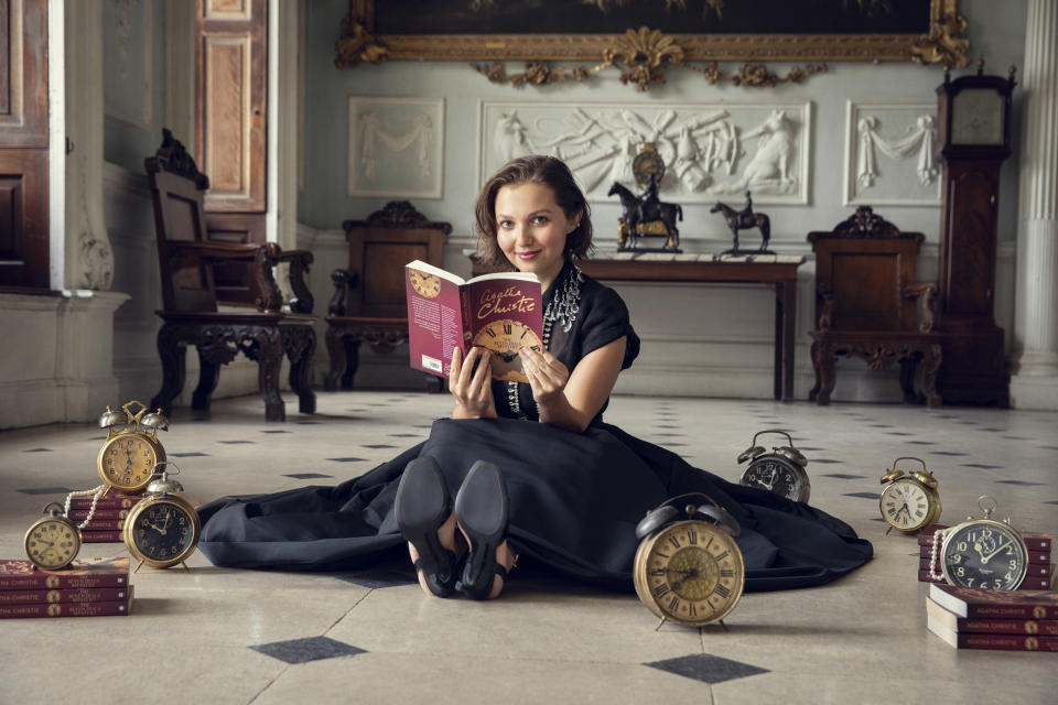 Mia McKenna-Bruce sitting on the floor in a black dress while reading The Seven Dials Mystery and surrounded by clocks