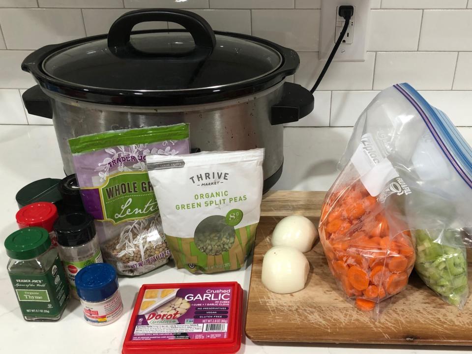 A Crock-Pot, seasonings, a container of crushed garlic, bags of lentils and split peas, and a cutting board on a white counter. On the cutting board is an onion sliced in half, a bag of chopped carrots, a bag of celery, and a knife.