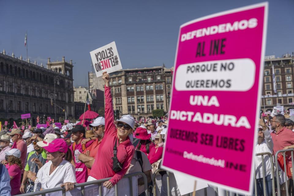 A person wearing a pink long-sleeved shirt and standing in a crowd holds up a sign near a larger poster in pink