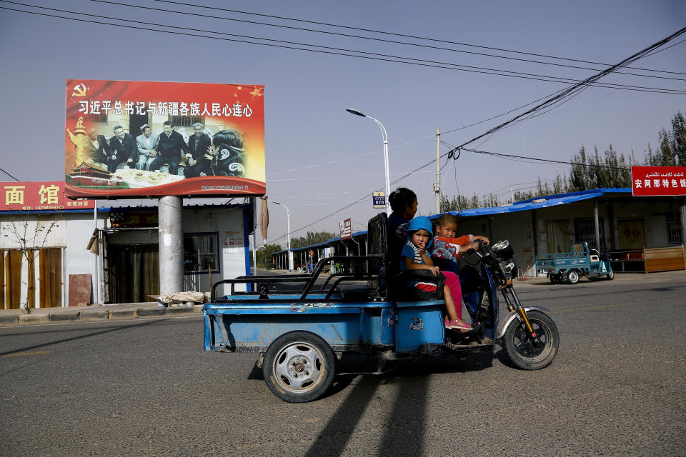 FILE - In this Sept. 20, 2018, file photo, an Uighur woman shuttles school children on an electric scooter as they ride past a propaganda poster showing China's President Xi Jinping joining hands with a group of Uighur elders in Hotan, in western China's Xinjiang region. The prominent British human rights lawyer Geoffrey Nice is convening an independent tribunal in London with public hearings in 2021, to look into the Chinese government's alleged rights abuses against the Uighur Muslim minority in the far western province of Xinjiang.(AP Photo/Andy Wong, File)