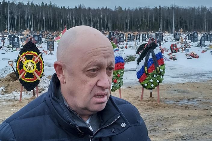Yevgeny Prigozhin, bare-headed, in a cemetery set with large floral wreaths, two red, white and blue, and one scarlet and yellow, with a star at its center.