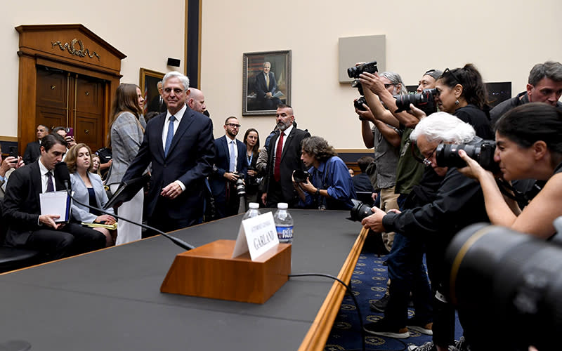 Attorney General Merrick Garland arrives for a House Oversight and Accountability Committee hearing. The photo is taken from an angle looking down the length of the table. On the left, Garland is walking toward his seat. On the left is a group of photographers taking pictures.