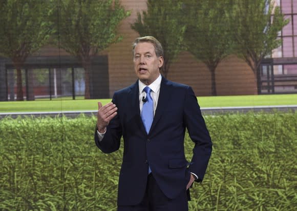 Bill Ford is shown speaking onstage, before an image of a green lawn in front of a brick building.