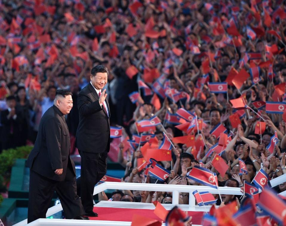 Xi Jinping and Kim Jong Un waving to crowds with North Korean and Chinese flags.