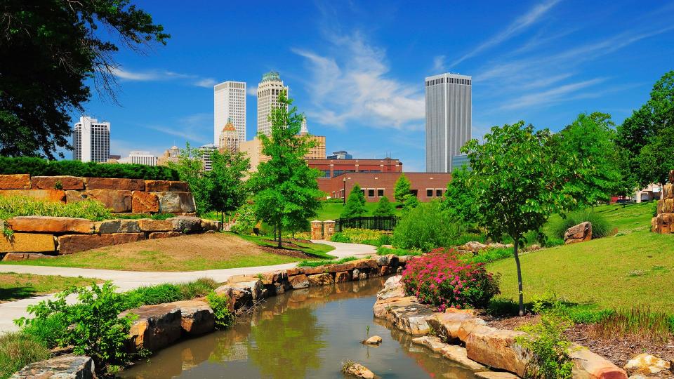 "Tulsa downtown skyline from a park with trees, grass, rocks, and a stream in the foreground.