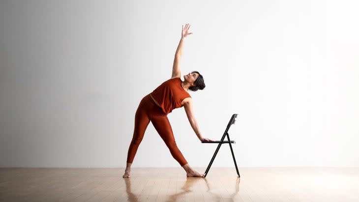 A woman with dark hair practices Triangle Pose with a chair for support. She is wearing copper colored clothes. The folding chair is black, the floor is light wood, the wall behind her is white