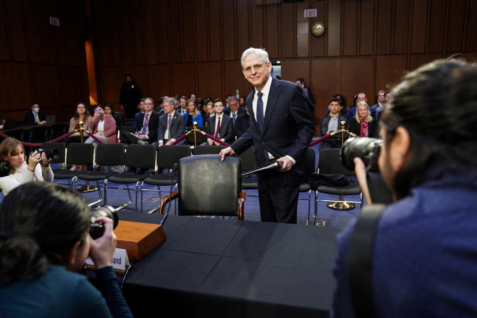 Merrick Garland stands next to an empty chair facing photographers in a wood-paneled room full of a few dozen seated people.