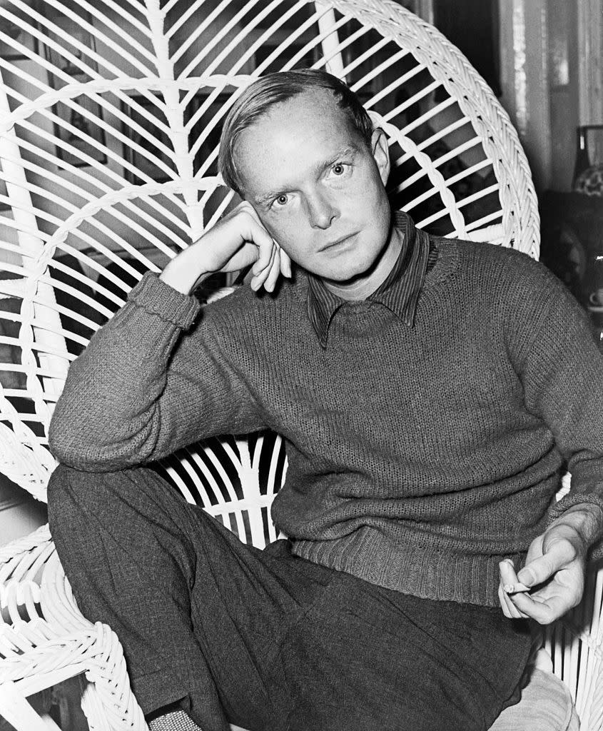 truman capote sits in a highbacked wicked chair and looks at the camera as he rests his head on one hand, he wears a sweater over a collared shirt and slacks