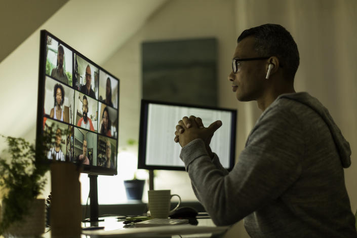 A person sits at a desk in a home office while other people are seen on a video conference on a monitor.