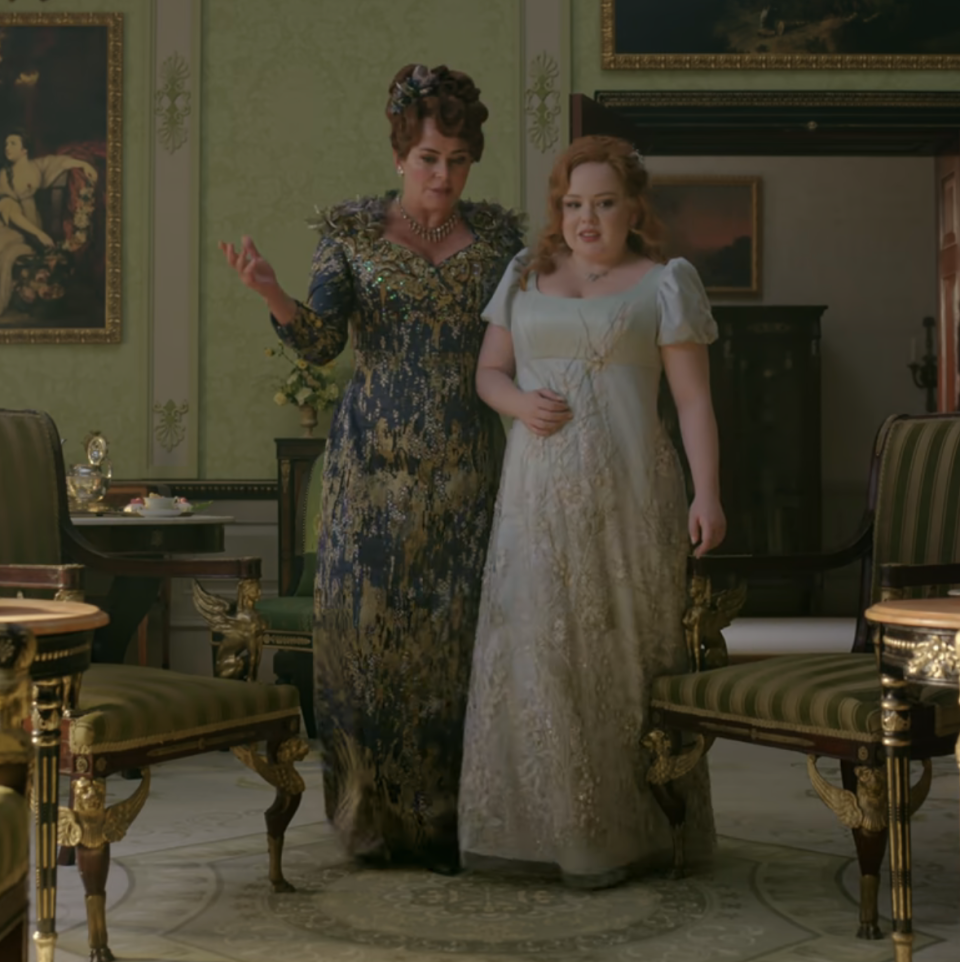Lady Featherington in a detailed gown walks with a pensive-looking Penelope, wearing an elaborate period dress, in an ornate room, from a scene in Bridgerton