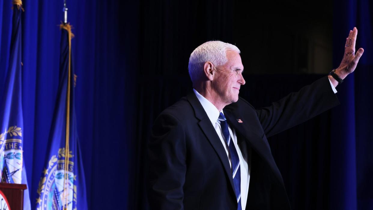 mike pence stands and waves, he faces to the right and wears a dark suit jacket, white collared shirt, and blue tie with a blue and white stripe