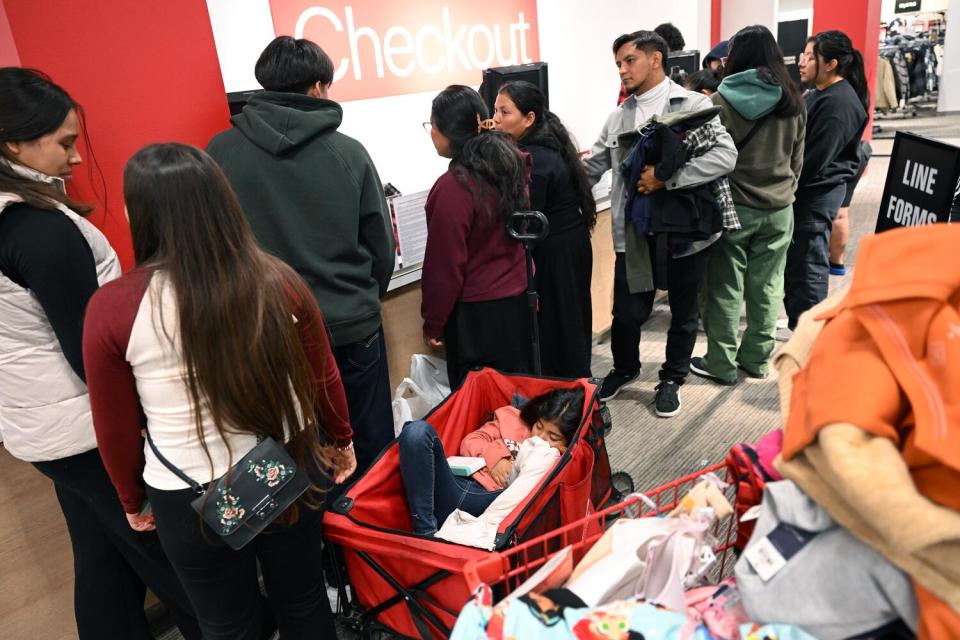 A line of people waiting at a sign reading "Checkout" at a store as a girl curls up in one family's cart for a rest.