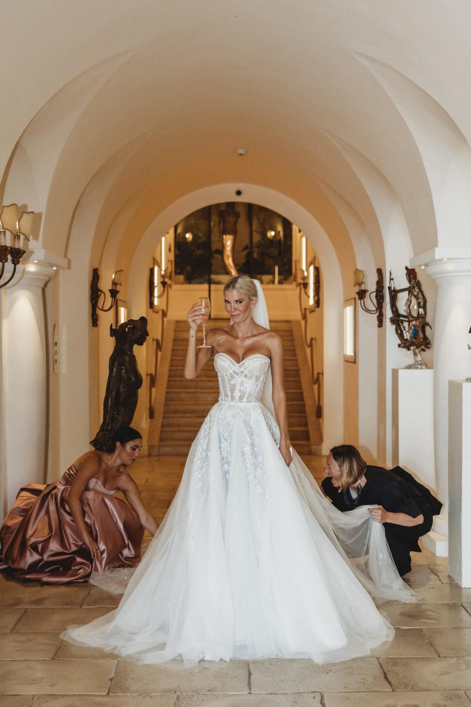 A bride raises a glass of champagne as two women adjust her train.