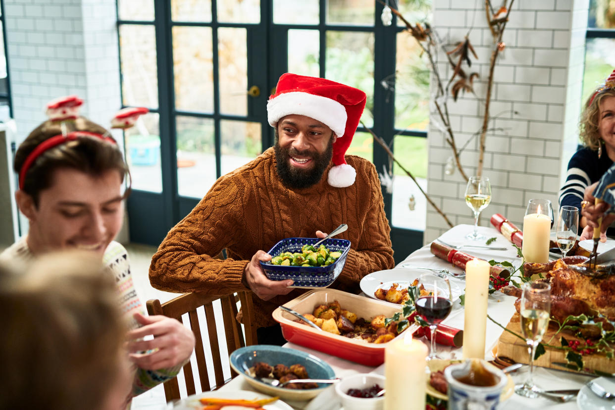 Mid adult black man with beard smiling and holding serving dish with sprouts, table set with roast dinner, traditional family Christmas at home