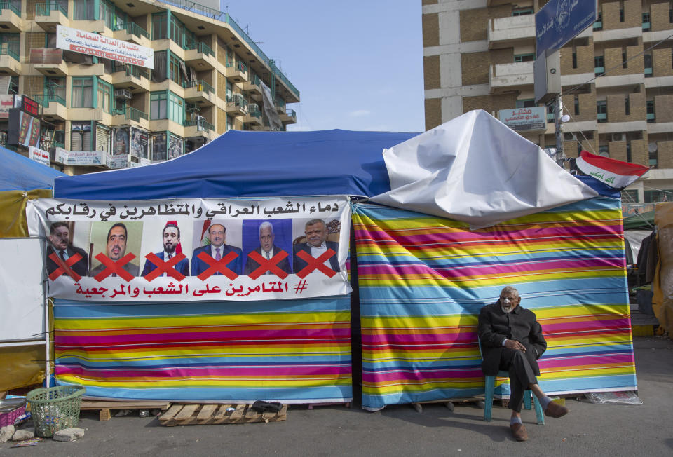 A protester sits next to a poster with defaced pictures of Iraqi politicians and Arabic that reads, "the blood of the protesters and the Iraqi people is on your necks, the conspirators on the people and the religious authority," during ongoing protests, in Tahrir square, in Baghdad, Iraq, Wednesday, Dec. 25, 2019. (AP Photo/Nasser Nasser)