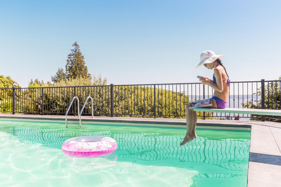 A teen sits on a diving board playing on her phone. 