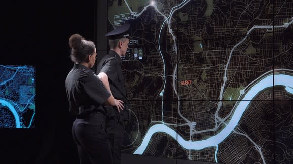 A police coordination team and officers discuss response tactics in front of large live screens in a modern office.