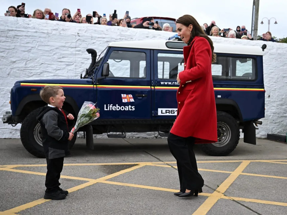 Kate Middleton and a little boy holding flowers smile at each other as Prince William walks towards them.