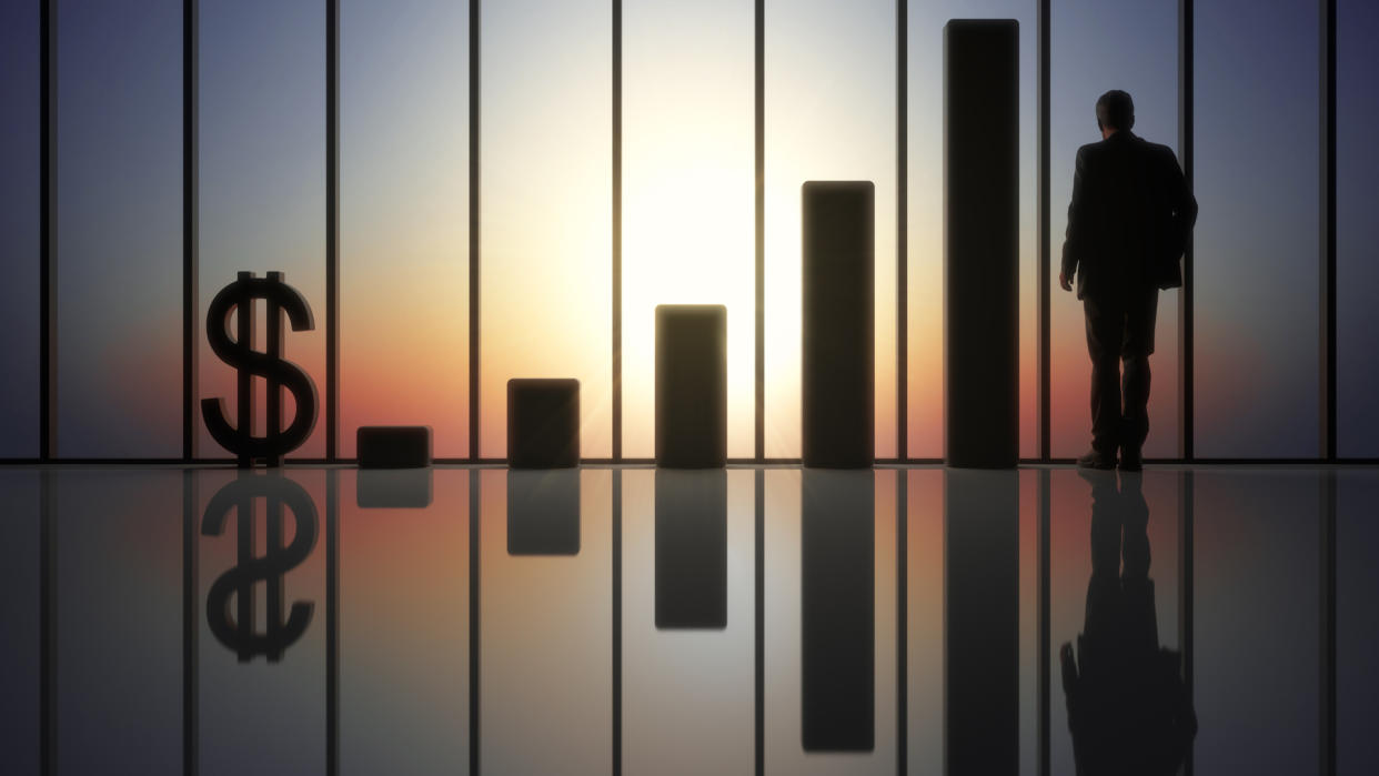 Suited businessman standing at panoramic window in futuristic hall with growing statistics and dollar sign at his left.