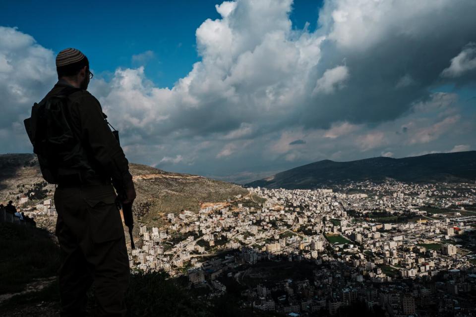 A man in a yarmulke holding a long gun and looking down from a hill to a city below