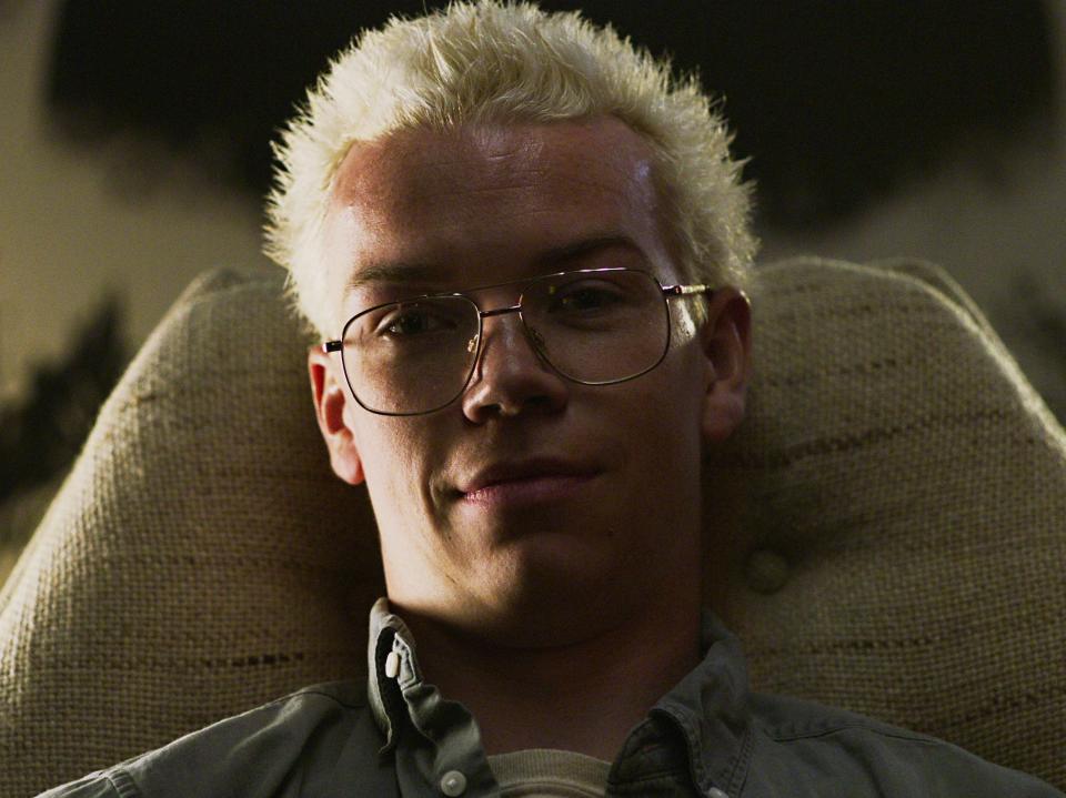 a man sitting back in a chair in black mirror bandersnatch. he has spiky, platinum blonde hair, large wire-rim glasses, and a smirk on his face. his face is lit only on one side, as if there's light coming in through a window