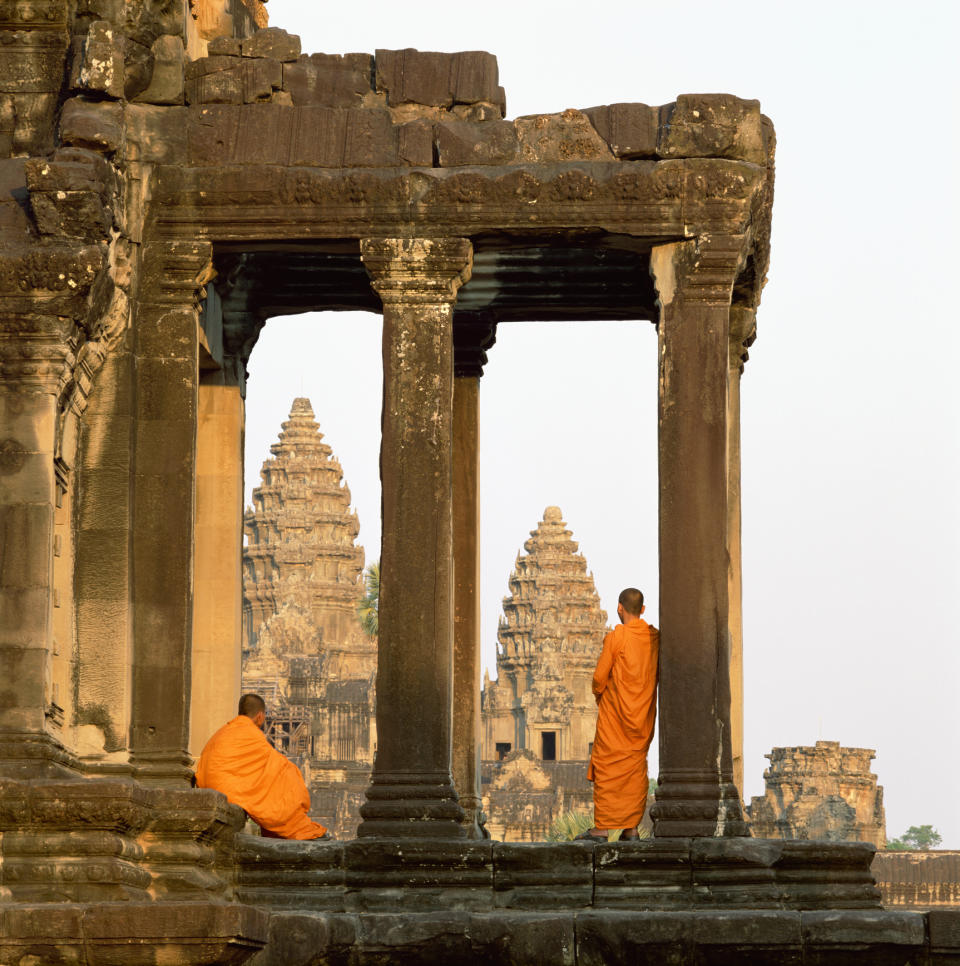 Two monks stand by a temple's ancient ruins, looking toward central towers