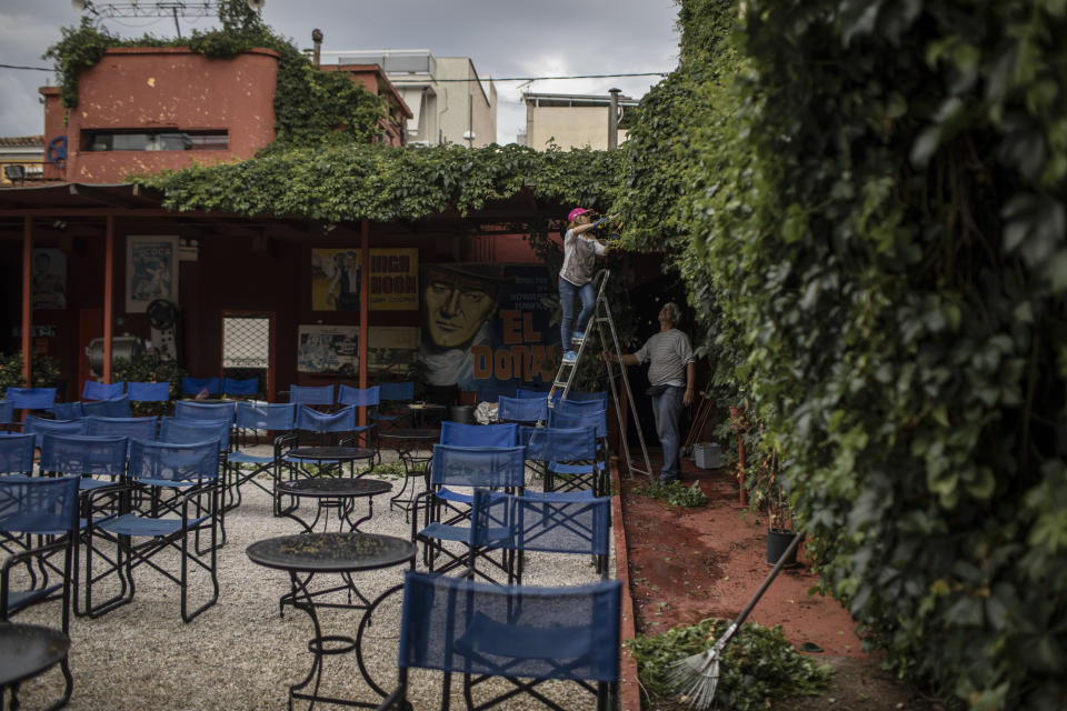 In this Friday May 29, 2020 photo Christina Tsouta, left, cuts leaves as projector operator Pavlos Lepeniotis holds the ladder at the Zephyros open-air cinema that specializes in films from past decades in the Petralona district in central Athens. (AP Photo/Petros Giannakouris)