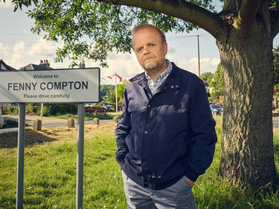 Toby Jones stands on a grassy piece of land next to a sign that reads 'Welcome to Fenny Compton'.