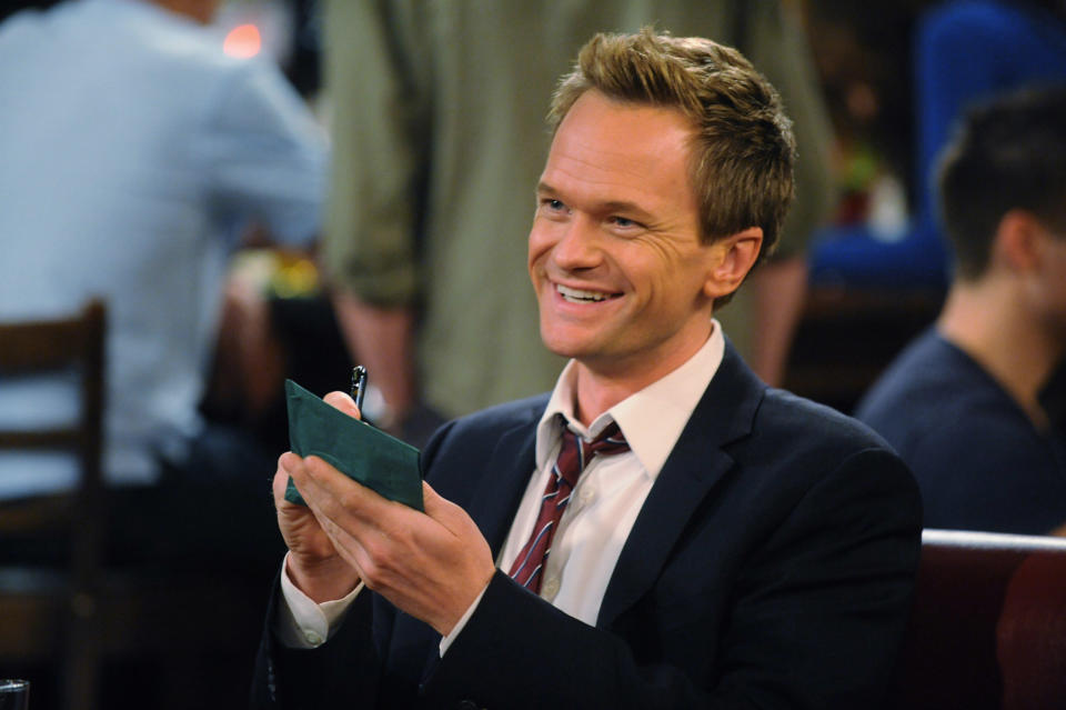 Barney Stinson from "How I Met Your Mother" smiles, holding the Playbook in a bar scene